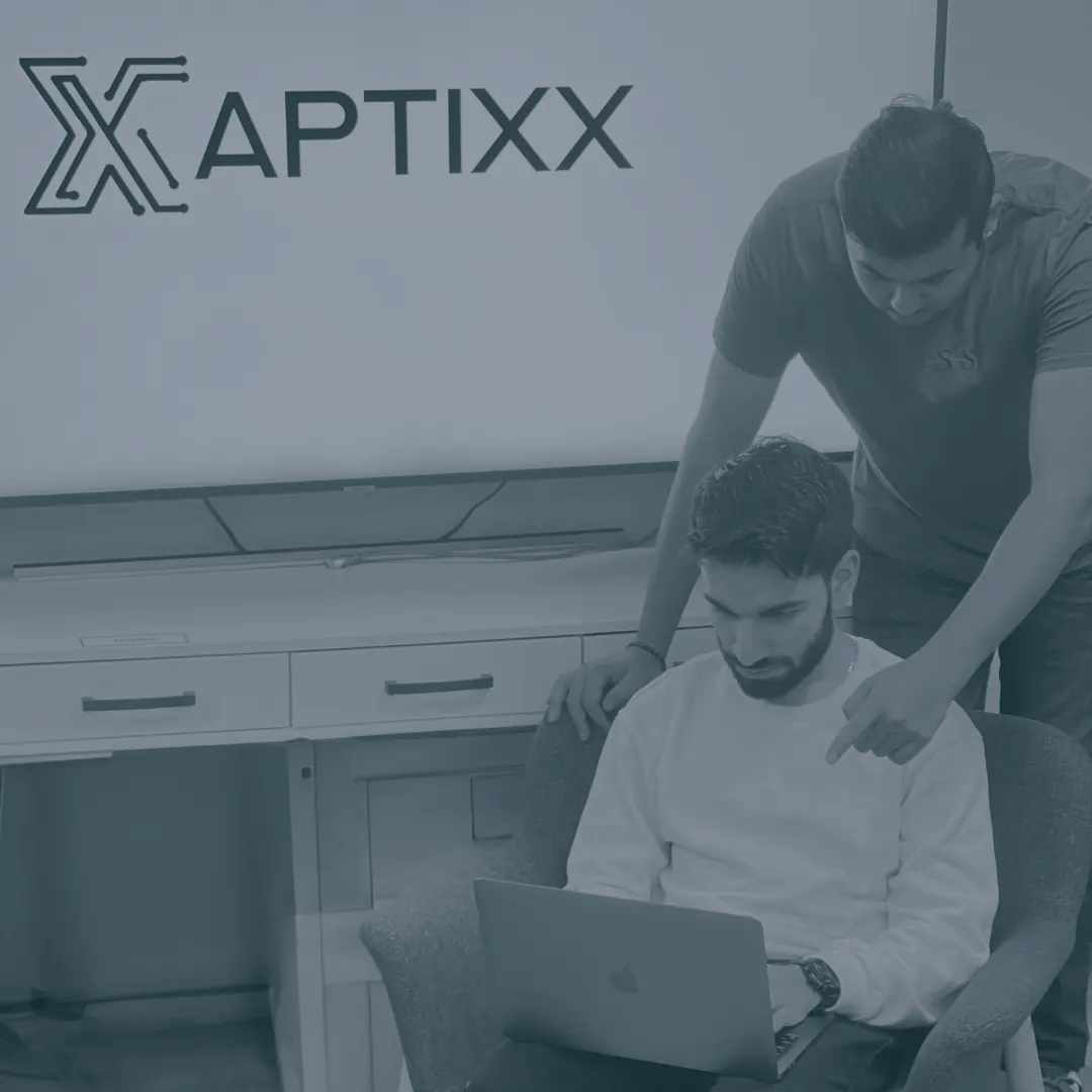 Two Aptixx engineers working together at a desk beneath a wall display carrying the Aptixx X mark